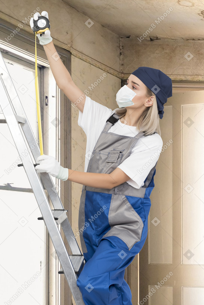 A woman in face mask standing with measuring tape on a ladder in a room