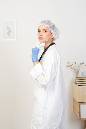 Vista lateral de una joven doctora poniéndose una máscara y mirando a la cámara