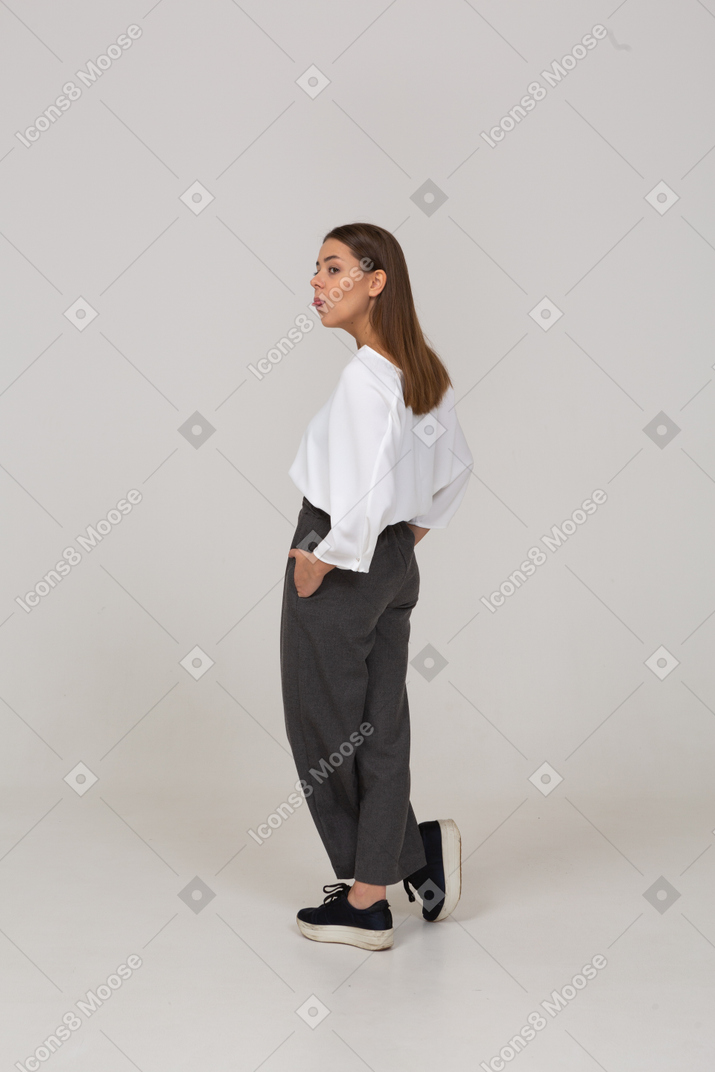 Vista traseira a três quartos de uma jovem com roupa de escritório, colocando as mãos nos bolsos e mostrando a língua