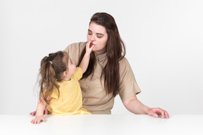 Kid girl sitting at the table and holding his mother nose