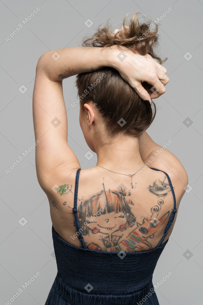Женщина стоит спиной к камере и поднимает волосы, чтобы показать татуированную спину