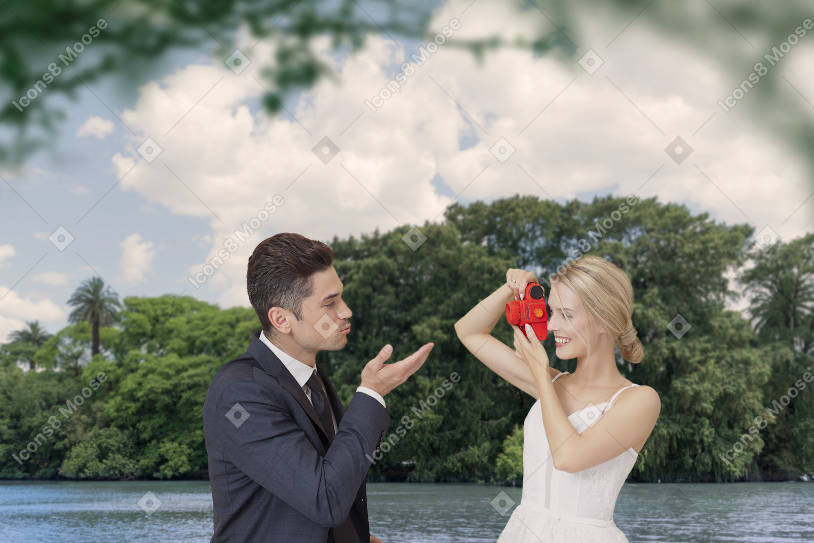 Uma noiva feliz tirando foto do noivo mandando um beijo para ela sob as árvores verdes perto do rio