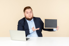 Junger übergewichtiger mann, der am schreibtisch sitzt und auf kleine tafel zeigt