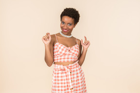 Young black short-haired woman in a checkered top and a skirt, posing against a plain peachy background