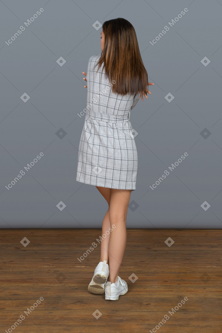 Unrecognizable slim woman with long brown hair standing back to camera and embracing herself