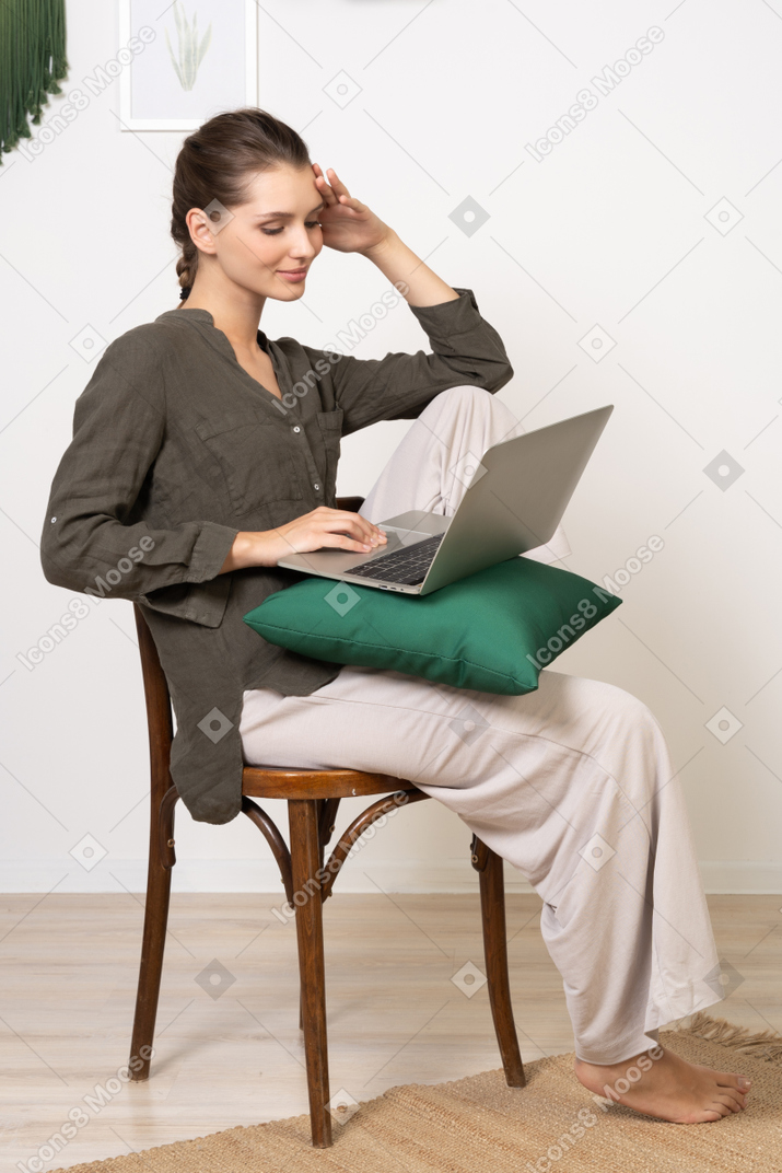 一位穿着家居服的年轻女子坐在椅子上，拿着笔记本电脑，摸着额头的四分之三视图