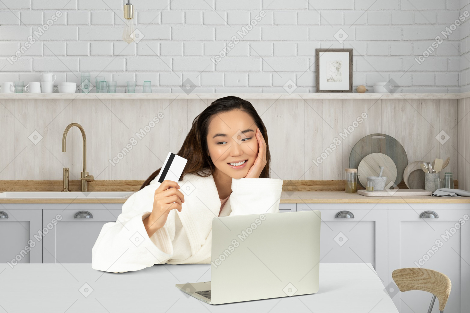 A woman sitting at a kitchen table holding a credit card and looking at a laptop
