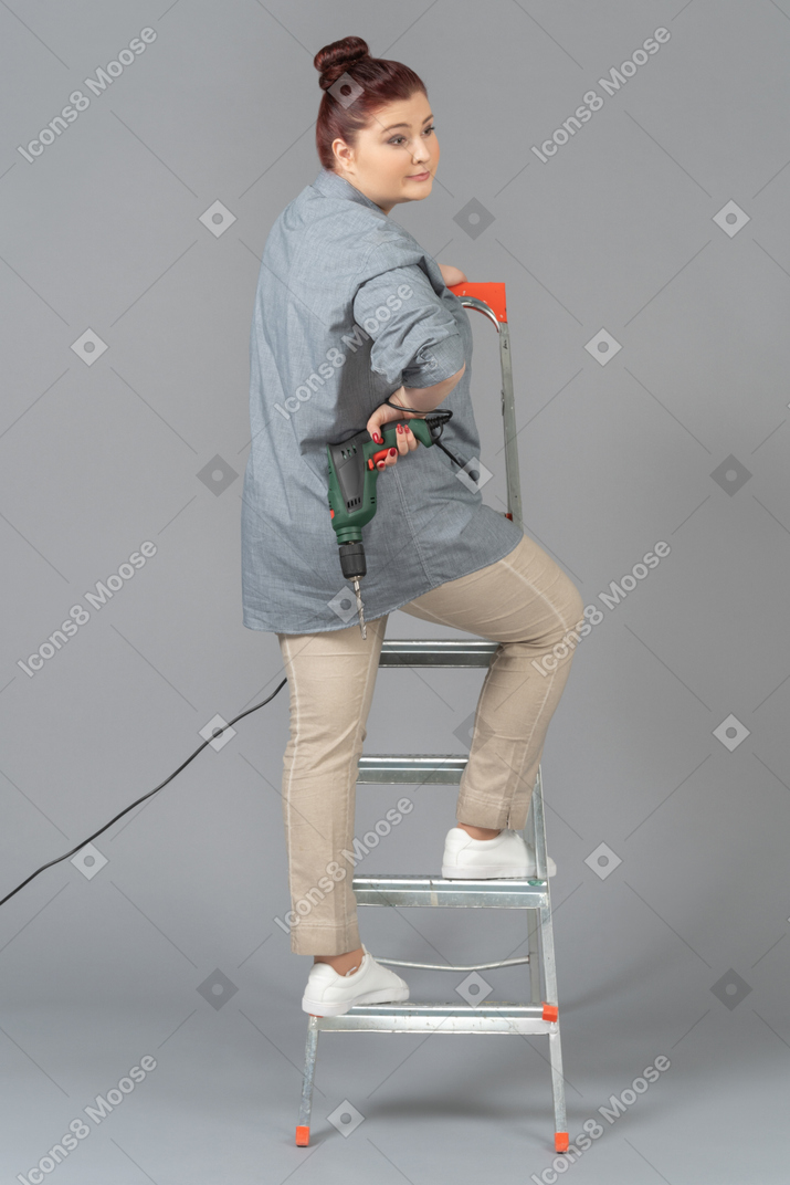 Woman standing half-way round on stepladder with a drill