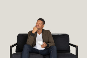 一个体贴的年轻男子坐在沙发上，端着一杯咖啡的前视图