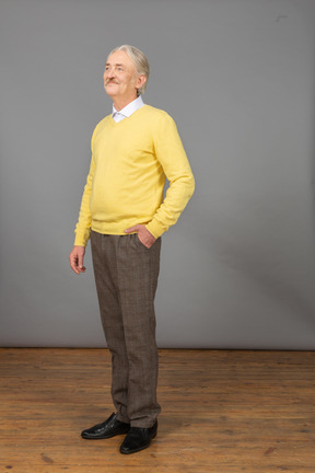 Three-quarter view of a pleased old man in a yellow pullover putting hand in pocket and looking aside