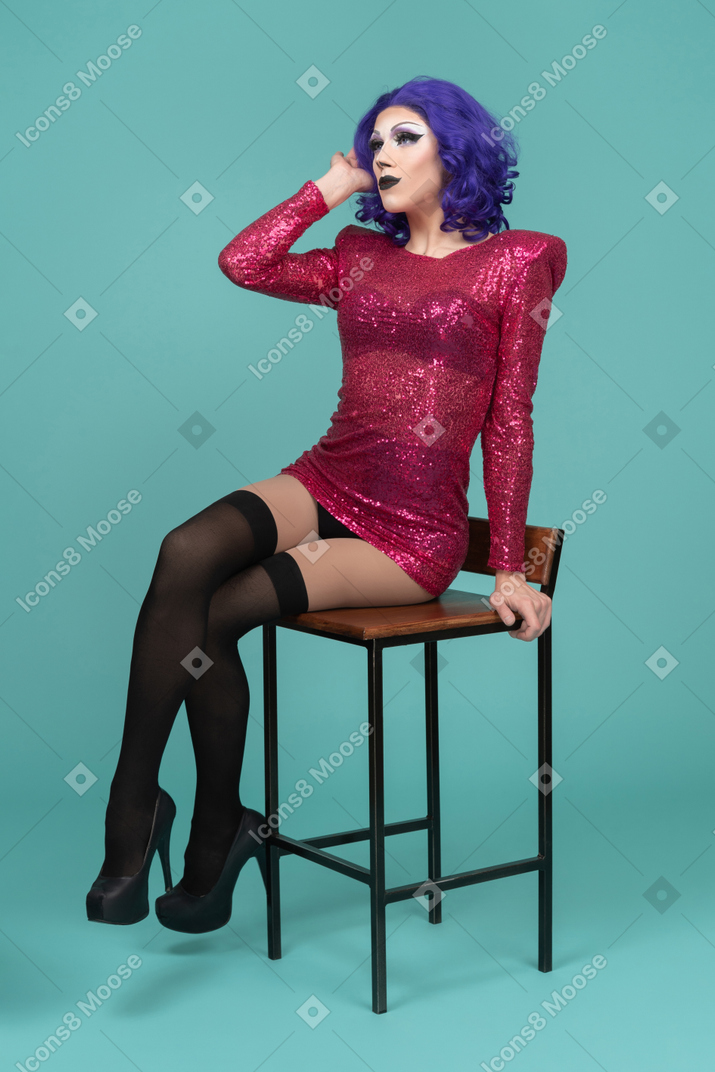 Трансвестит в розовом платье поднимает руку к лицу, сидя на стуле