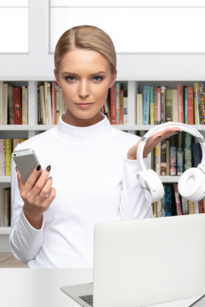 A woman holding a cell phone and headphones in front of a laptop