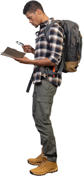Vista lateral de un turista con una mochila examinando el bloc de notas a través de una lupa