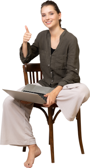 Front view of a smiling young woman sitting on a chair with a laptop & showing thumb up