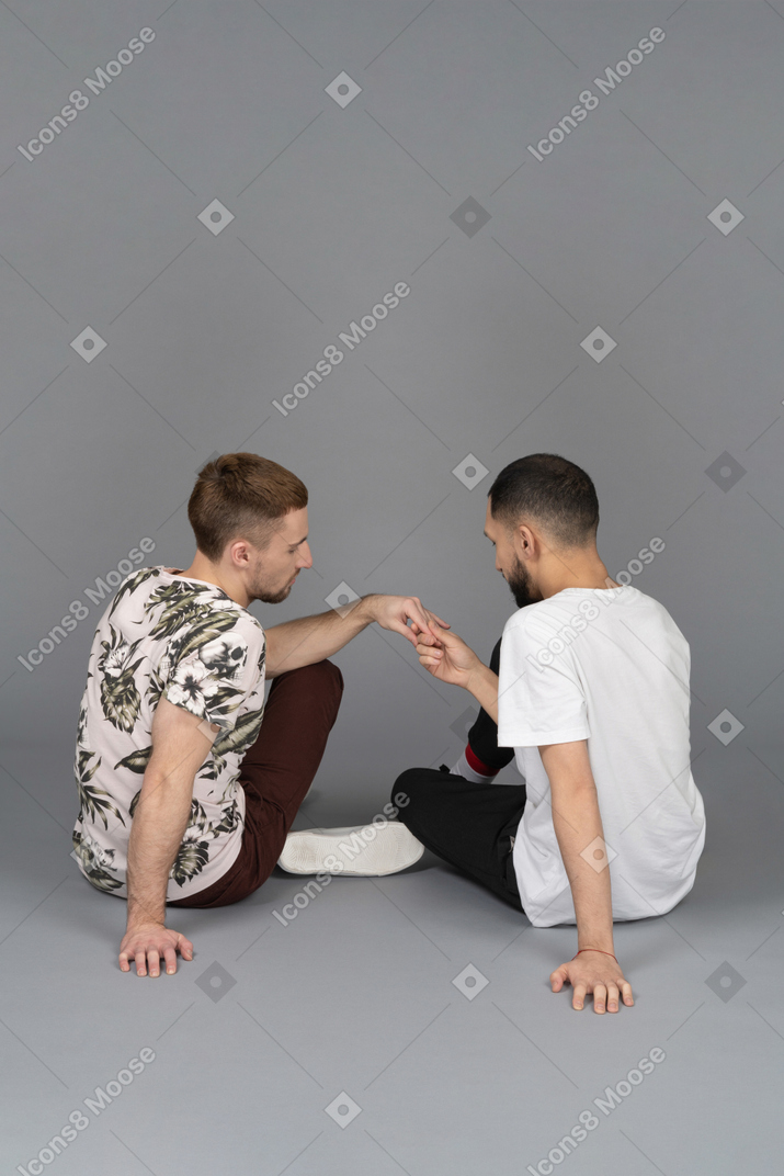 Vista posteriore di due giovani uomini seduti sul pavimento e tenendosi per mano