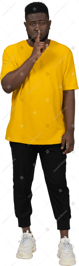 Vista frontal de un joven de piel oscura con camiseta amarilla que muestra el gesto de silencio