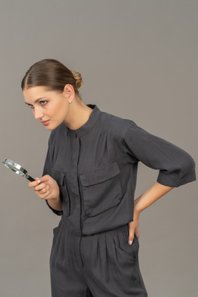 Three-quarter view of a young woman in a jumpsuit holding a magnifying glass