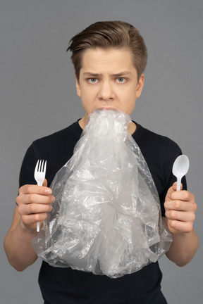 Close-up male holding plastic fork and spoon with plastic wrap in his mouth
