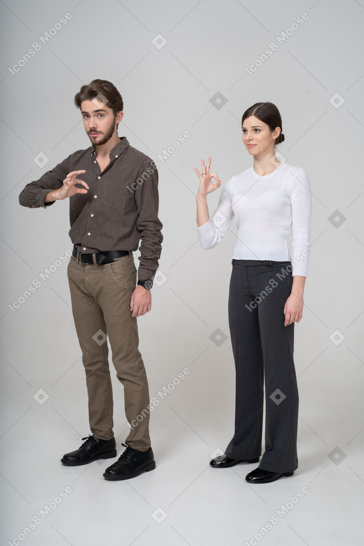 Vista lateral de um jovem casal com roupa de escritório mostrando sinal de ok
