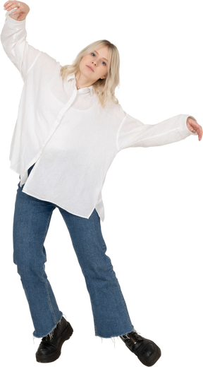 Front view of a blonde female in casual clothes dancing on her tiptoes and raising hands looking at camera
