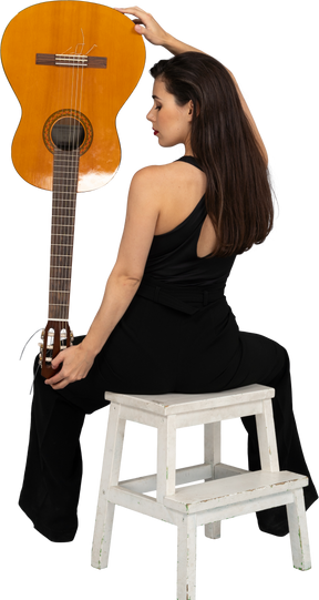 Rückansicht einer jungen dame im schwarzen anzug, die die gitarre kopfüber hält und auf hocker sitzt