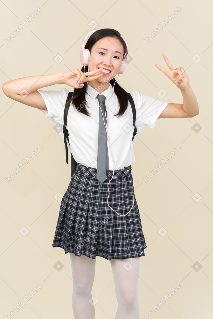 Asian school girl listening to music in headphones and showing v sign