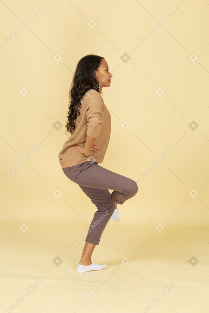 一位黑皮肤的年轻女性，抬高腿＆把手放在臀部上的侧视图
