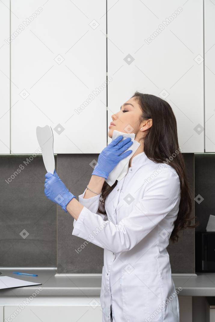 Side view of a female doctor holding a mirror and wiping her face