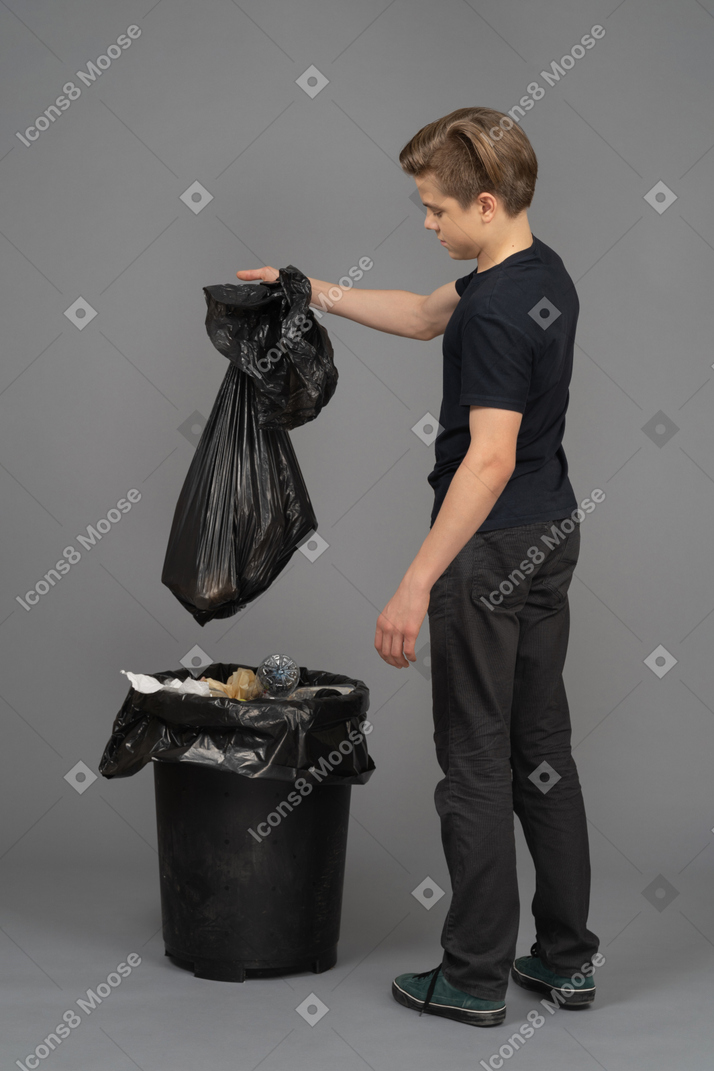 A young man holding a trash bag above a waste bin