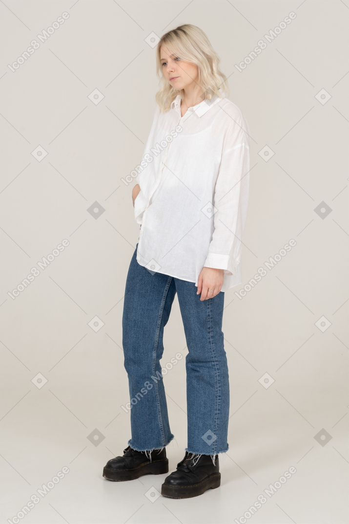 Three-quarter view of a blonde female in casual clothes putting hand in pocket and looking down sadly
