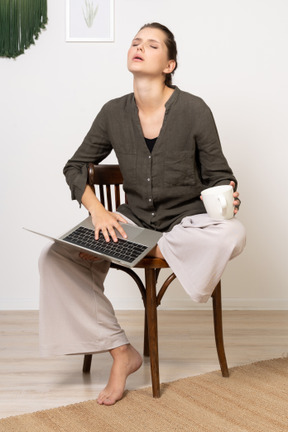 Vorderansicht einer müden jungen frau, die hauskleidung trägt und auf einem stuhl mit einem laptop und einer kaffeetasse sitzt