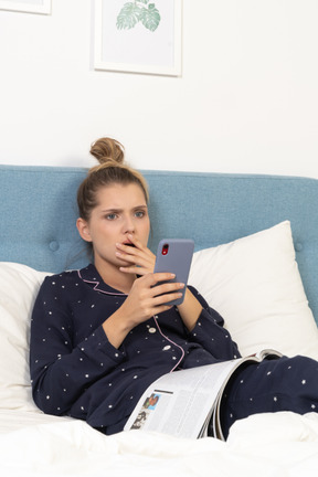 Close-up de uma jovem chocada de pijama, deitada na cama enquanto navega na internet