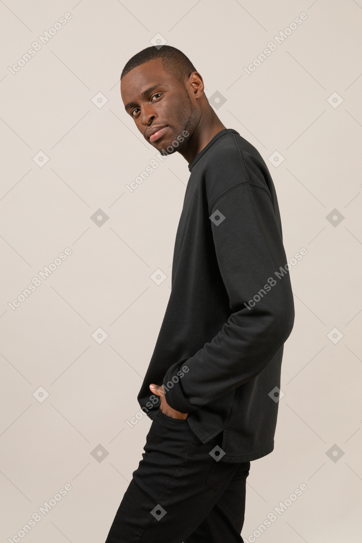 Side view of a young man keeping hands in pockets and looking at camera