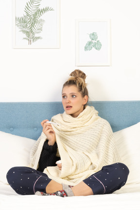 Front view of a perplexed young lady wrapped in white blanket sitting in bed with thermometer