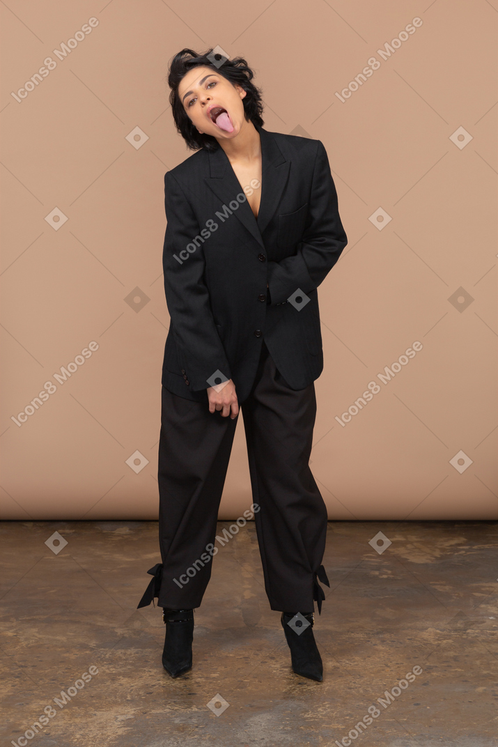 Vista frontal de una mujer de negocios en un traje negro inclinado hacia adelante y mostrando la lengua mientras mira a la cámara