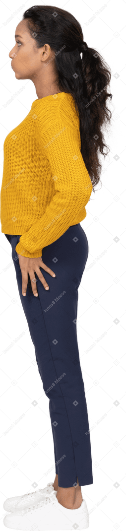 Vista lateral de una niña en ropa casual de pie con la mano en la cadera y haciendo muecas