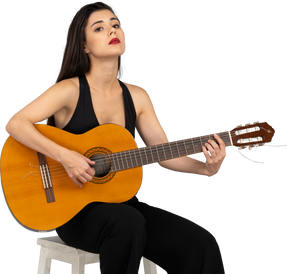 Front view of a sitting young lady in black suit holding the guitar and raising head