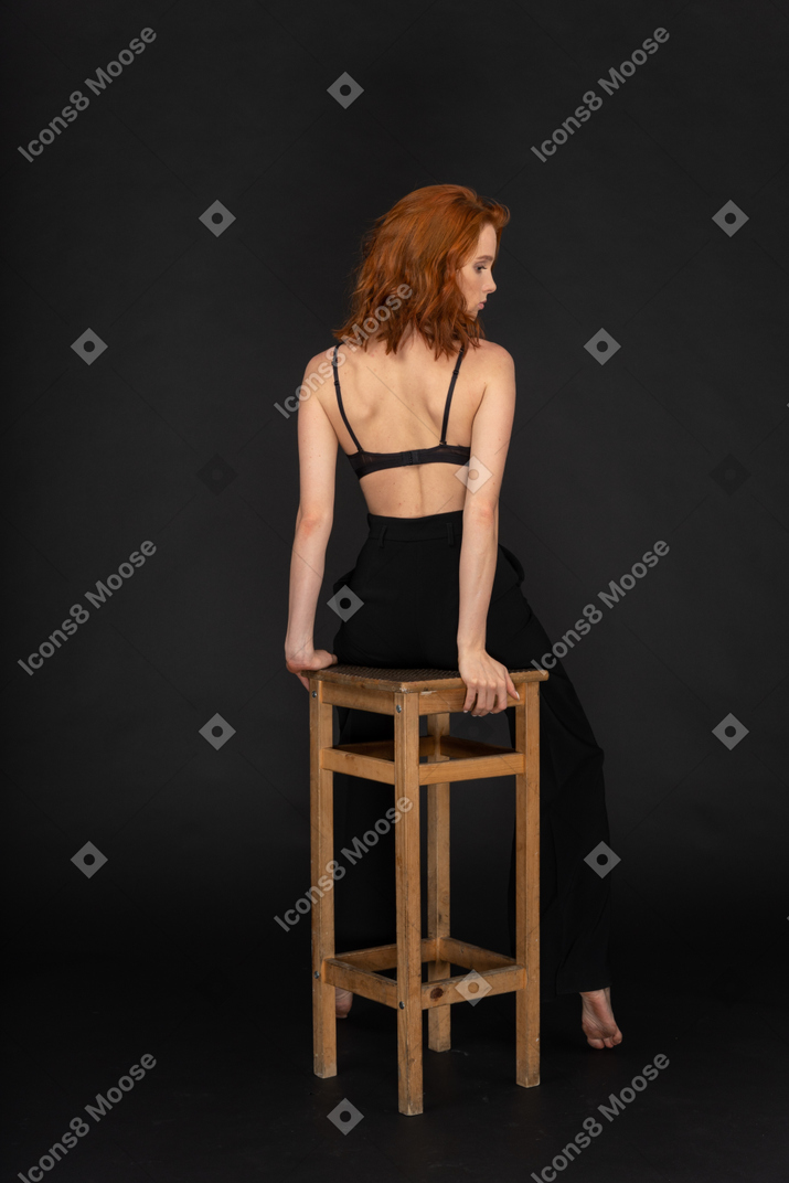 A back view of the beautiful woman dressed in black pants and bra, sitting on the wooden chair and looking to the right