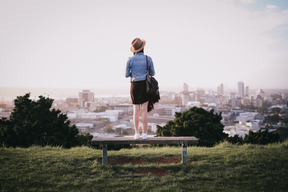 Молодая женщина стоит на скамейке и наслаждается видом на город