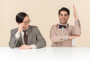 Dois jovens nerds sentado à mesa e um deles está levantando a mão