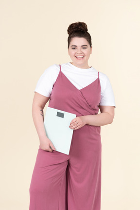 Young plus size woman in a pink jumpsuit, posing with scales against a pastel yellow background