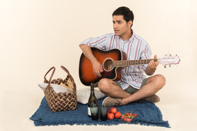 Young caucasian guy sitting near picnic basket on the blanket and playing guitar