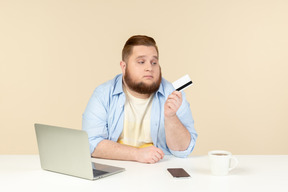 Trauriger schauender junger überladener mann, der am schreibtisch sitzt und bankkarte hält