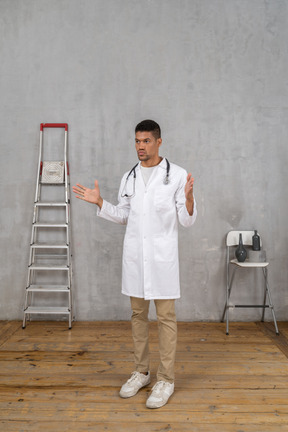 Vista de três quartos de um jovem médico gesticulando em uma sala com escada e cadeira