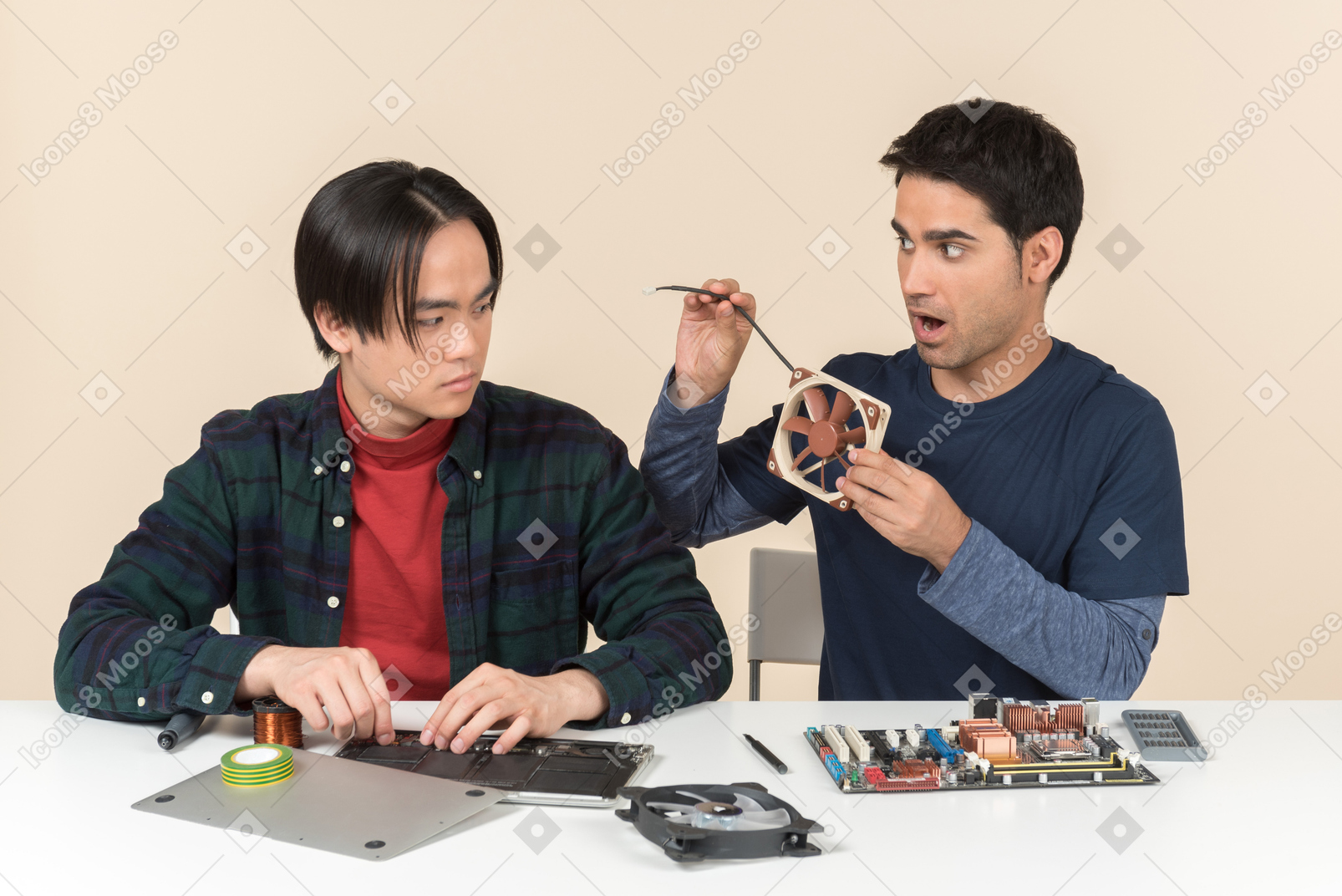 Two young geeks sitting at the table with details on it and one of them looking surprised