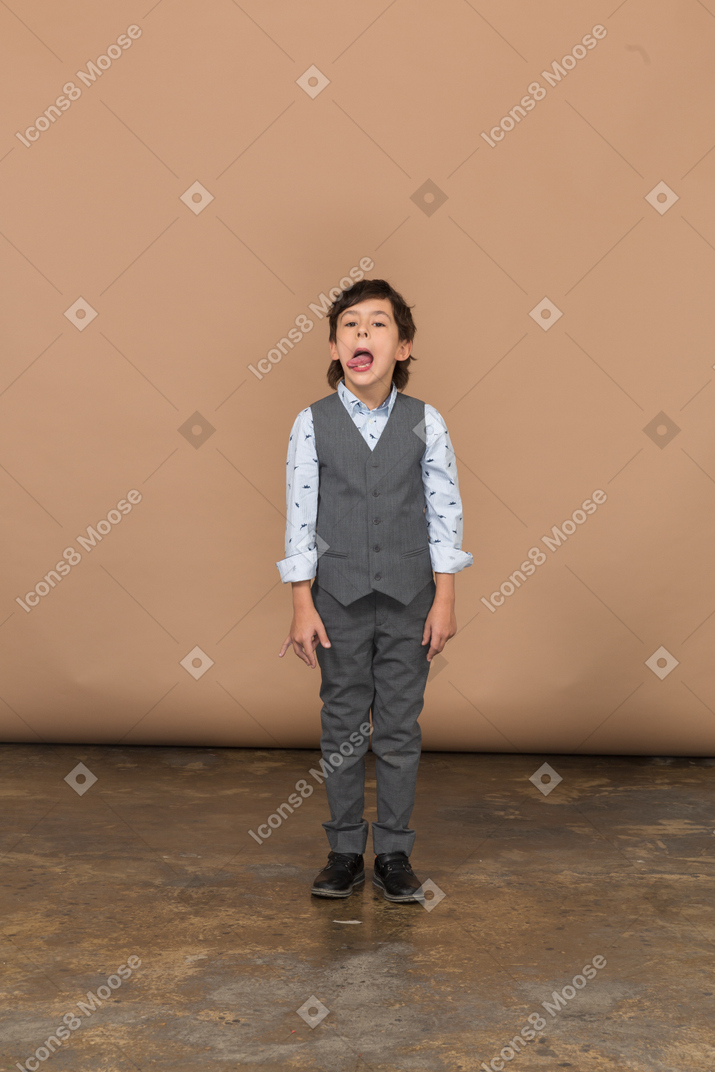 Vue de face d'un garçon mignon en costume gris faisant des grimaces et montrant la langue