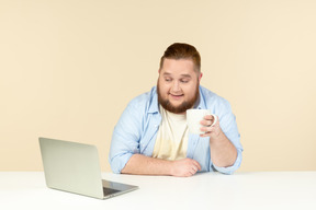 Junger übergewichtiger mann, der vor laptop sitzt und tee trinkt