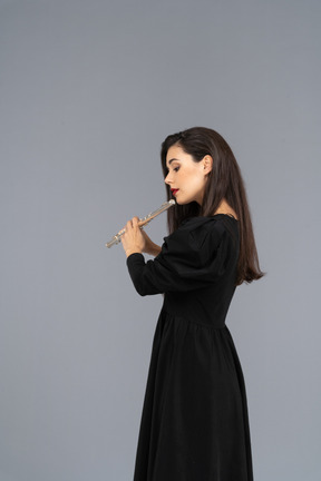 Vista lateral de uma jovem séria de vestido preto tocando flauta
