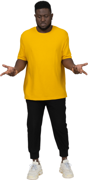 Vista frontal de un joven hombre de piel oscura gesticulando pensativo en camiseta amarilla