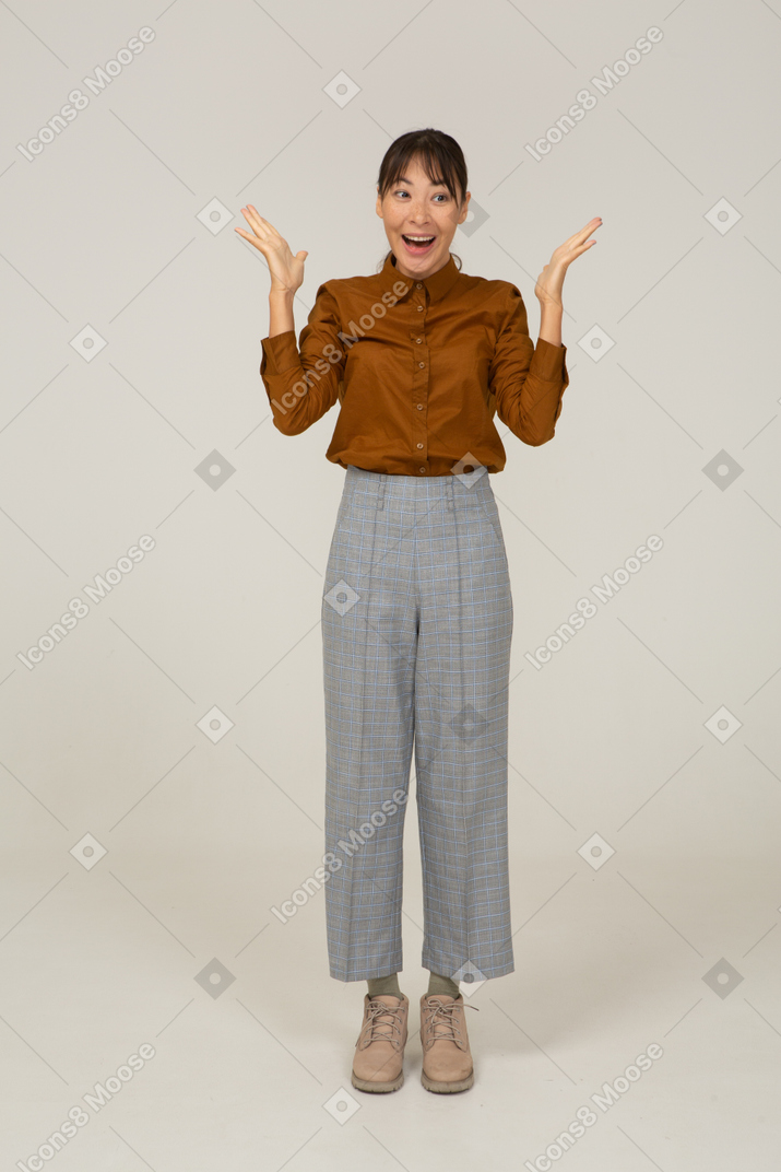 Front view of a surprised young asian female in breeches and blouse raising hands